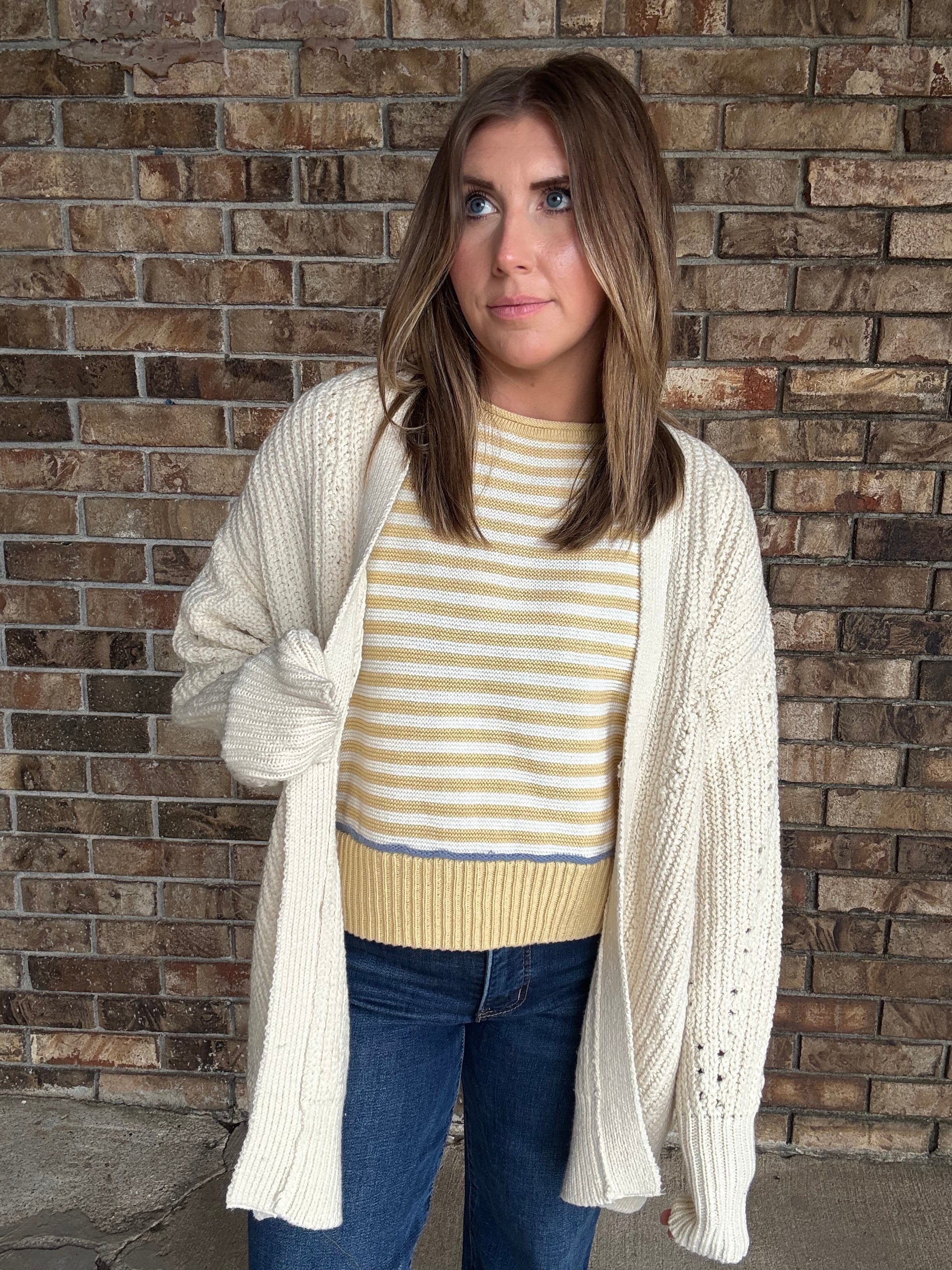 Woman wearing an oversized ivory open knit cardigan layered over a striped sweater and dark wash jeans