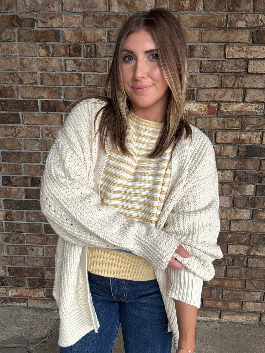 Woman wearing an oversized ivory open knit cardigan layered over a striped sweater and dark wash jeans