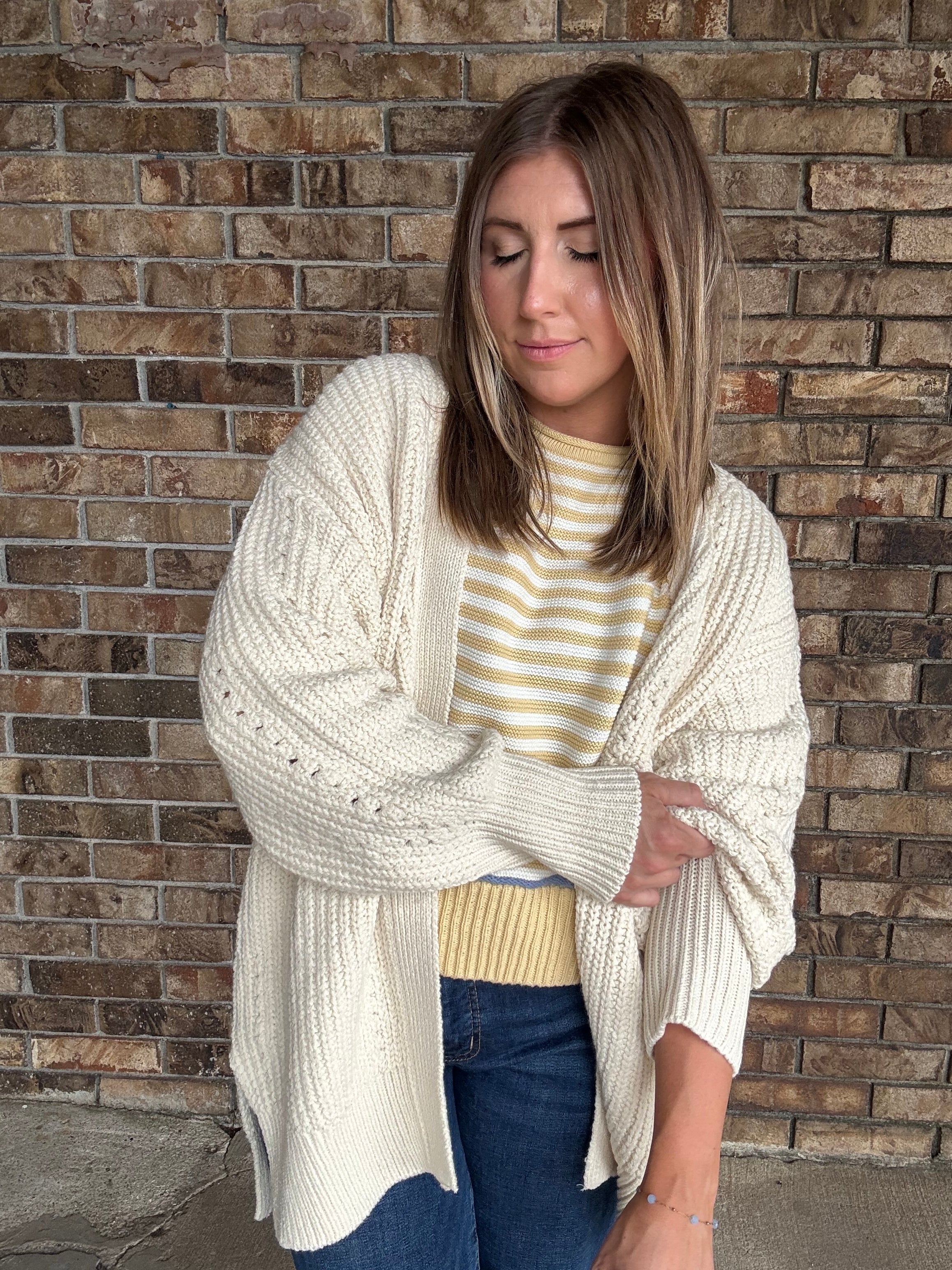 Woman wearing an oversized ivory open knit cardigan layered over a striped sweater and dark wash jeans