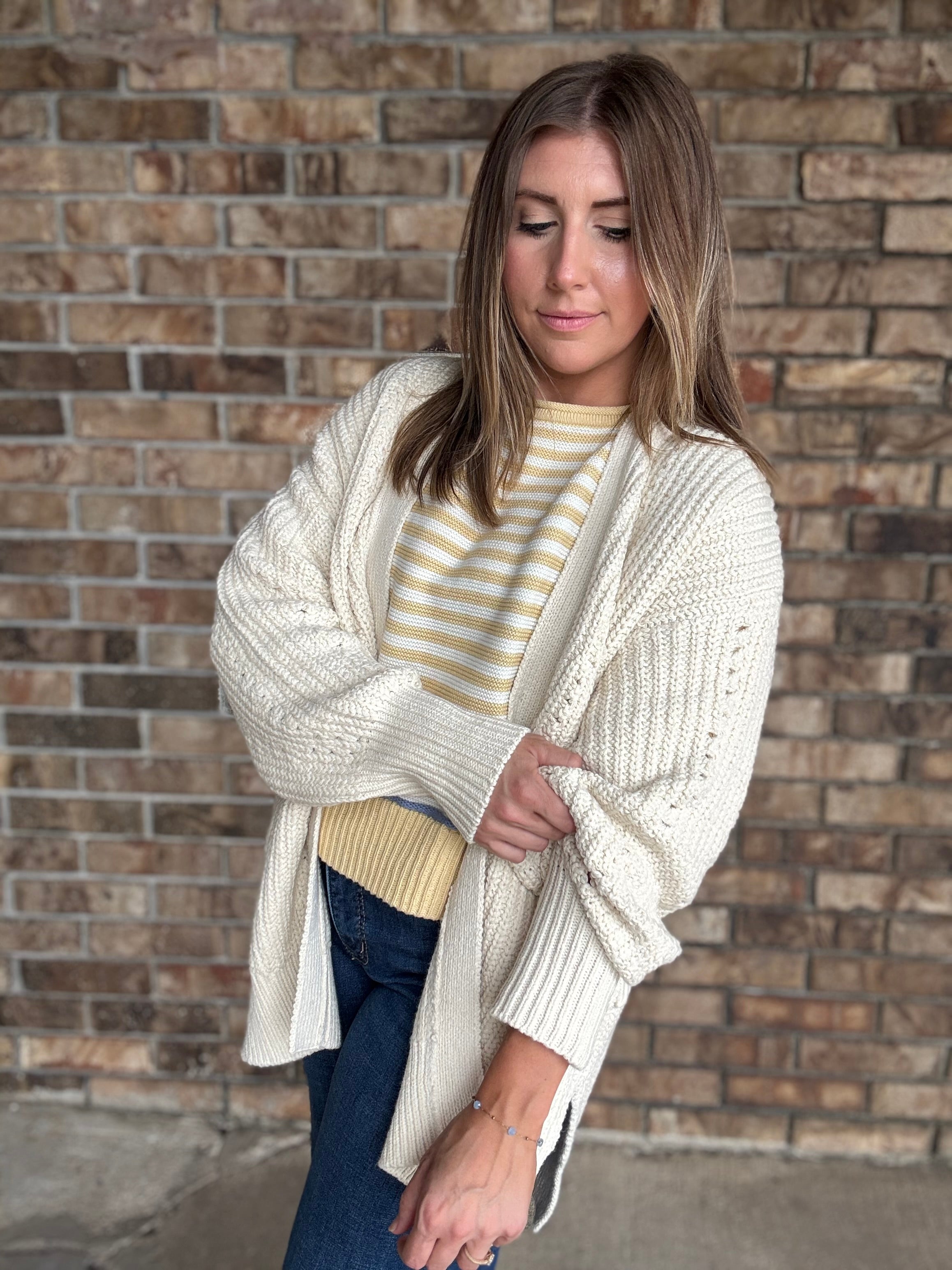 Woman wearing an oversized ivory open knit cardigan layered over a striped sweater and dark wash jeans