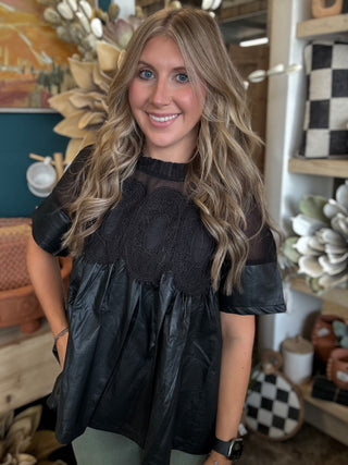 Woman wearing a black lace overlay faux leather babydoll top with sheer mesh neckline and short sleeves.